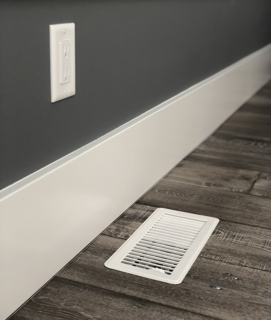 White floor vent next to wall with electrical socket. Wood flooring and white simple trim
